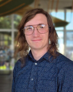 Aiden Wells with long brown hair and glasses wearing a dark patterned shirt, smiling in front of a blurred indoor setting