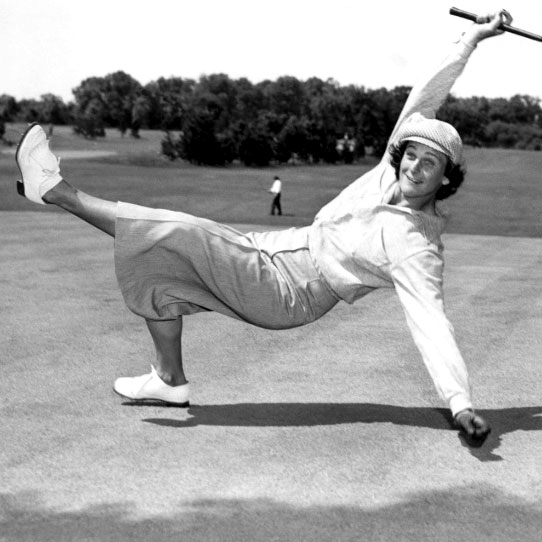 Female golfer, Babe Zaharias, kicks her foot in the air while holding a golf club