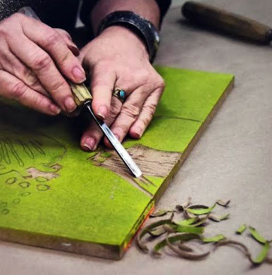 Two hands use a tool to carve away a block for printing