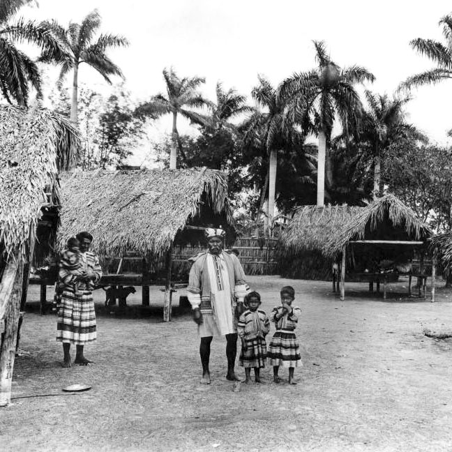 Seminole family stands in front of a collection of chickee huts, thatched A-frame shelters with pole supports