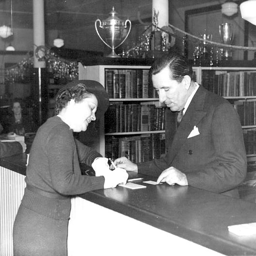 Old photo of a woman signing a guest book at a counter with a male clerk assisting