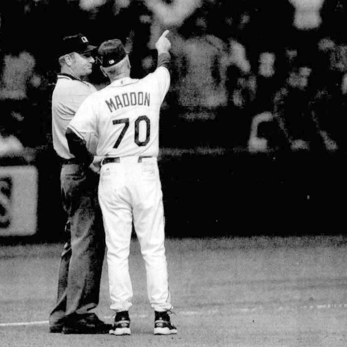 Baseball player Maddon stands in his uniform with his back to the photographer with another man pointing up, presumably at an instant reply video outside of view
