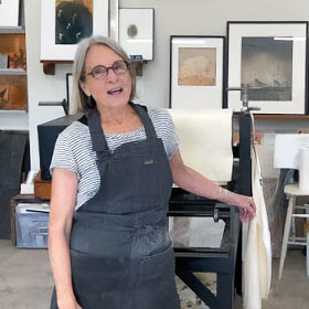 Artist Martha E Johnson stands next to a printing press wearing an apron and smiling