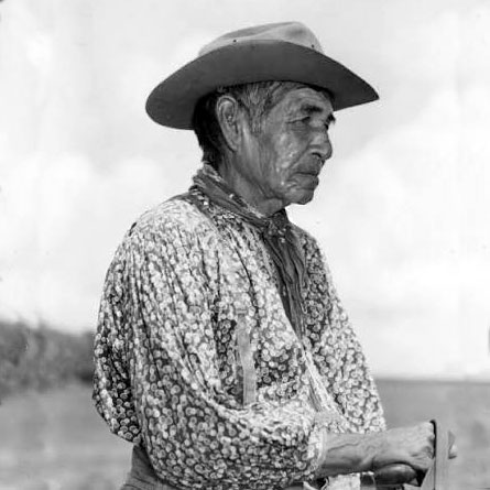 Black and white photo of a man wearing a cowboy hat and collared shirt with reins in his hand suggesting he is sitting on top of a horse