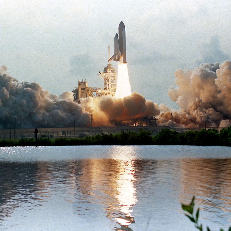 A space shuttle launching off the pad at what appears to be dusk with water in the foreground