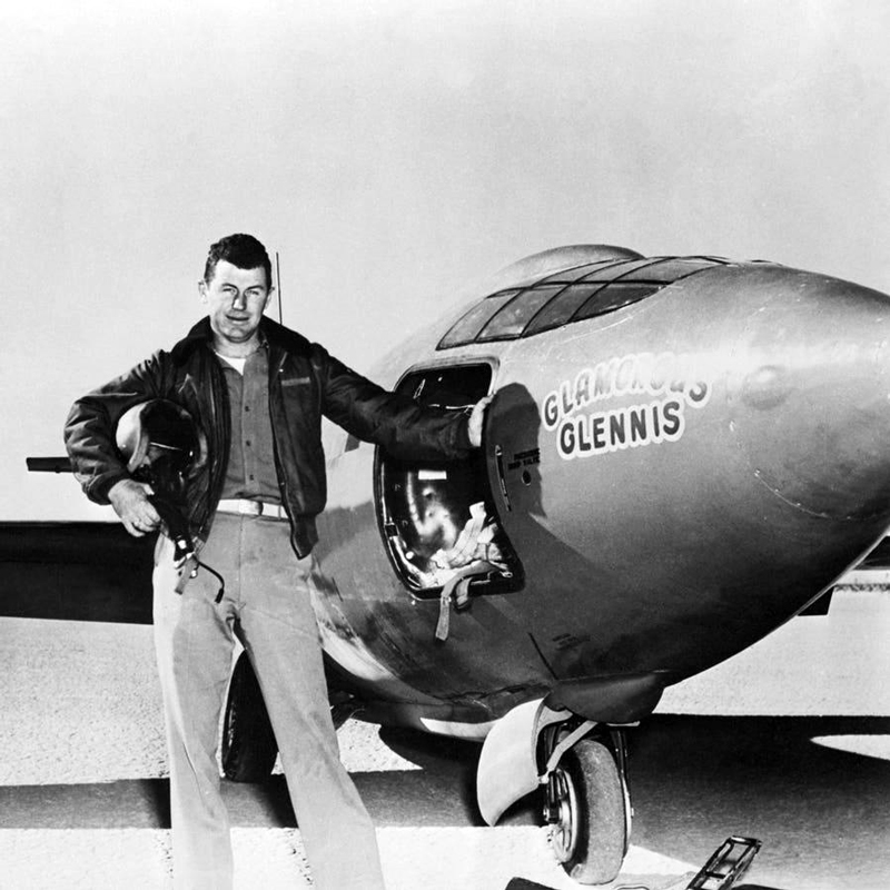 Pilot Chuck Yeager, a man with dark hair wearing a leather jacket, stands next to a plane named 