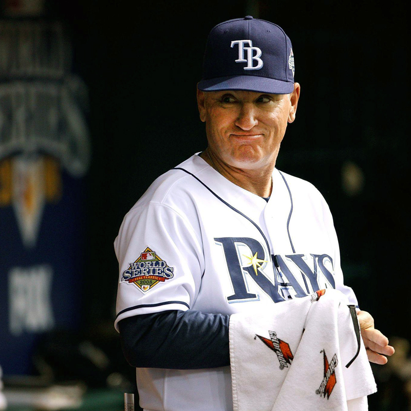 Joe Madden stands in front of a team banner wearing a Rays uniform while wiping his hands on a towel