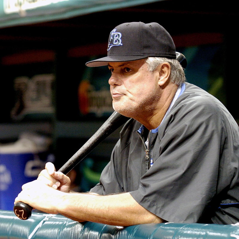 Manager Lou Piniella leans on a fence while holding a bat wearing a dark jersey and hat