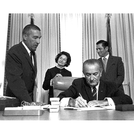 President Lyndon B. Johnson at a desk signing a document with staff behind him witnessing the act