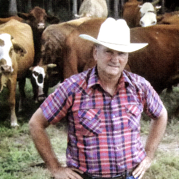 Photo of a rancher, JB Starkey, wearing a plaid shirt and cowboy hat, standing in front of a herd of cows