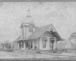 The Orange Belt Railway depot, which once stood at the corner of 1st Ave S and 2rd St. A narrow gauge engine is to the left of the platform, and the end of a boxcar is visible to the right. Four men and a boy stand on the platform. Orange Belt Investment Company office is in the background. c. 1889