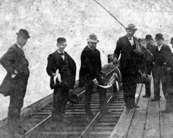Group of men in early 20th-century attire standing on a wooden dock or platform, some holding fish