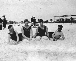 Bathers on St. Pete Beach. 24 February 1915. Four people in early 20th-century swimwear lying on a sandy beach, with other beachgoers and a pavilion structure in the background