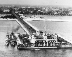 Aerial black-and-white photo of USS Northampton CG-13 docked at a pier with a large waterfront building, connected by a long causeway to shore