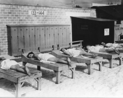 Sunbathers in the Solarium at the Spa on Spa Beach. c. 1933 People lying on wooden lounge beds in the sand near numbered lockers 133–144 at a beach facility, circa mid-20th century