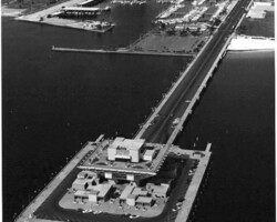 Aerial black-and-white photo of a long pier extending into the water, with a building complex at its end and a city skyline in the background