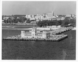 Aerial black-and-white photo of a waterfront pier with a large terminal building, parked cars, and a city skyline in the background