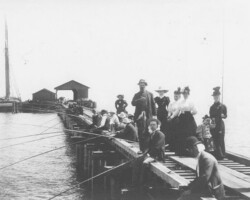 Historical black-and-white photo of a group of people fishing and gathering on a wooden pier, with a sailboat moored nearby