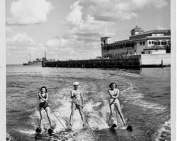 Three people water skiing in formation near a pier and large waterfront building, vintage black and white photo