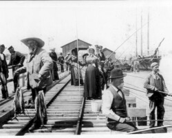 Historic black-and-white photo of men and women fishing from a wooden pier with a sailing ship docked in the background