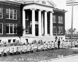 Children performing a flag drill in formation outside a brick school building, 1913