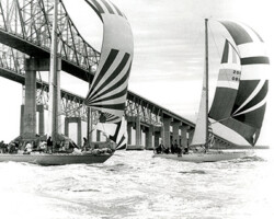 Sailboats with large spinnaker sails racing on the water beneath a steel bridge, black and white photo
