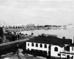 Historic black-and-white aerial view of a waterfront marina with docked boats, a boulevard, and white stucco buildings in the foreground