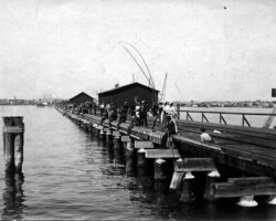 Historic black-and-white photo of people fishing from a wooden pier over calm water, with a waterfront building and distant shoreline in the background