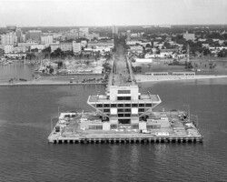 Aerial black-and-white view of a pier structure extending into the water, with a multi-story building at its end and a city skyline in the background