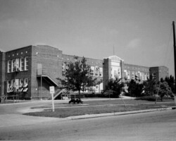 Black-and-white photo of a large brick school building with exterior staircases, trees, and a grassy traffic circle in the foreground
