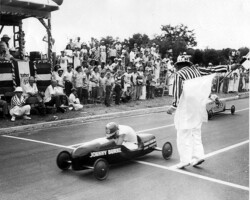 Vintage soap box derby race with a child driving a car labeled "Johnny Burke" crossing the finish line before a crowd of spectators