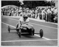 A young Johnny Burke wearing goggles and a helmet races a soap box derby car down a track before a large crowd of spectators