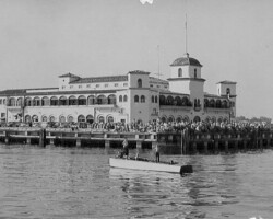 Historic black-and-white photo of a large Spanish-style waterfront building on a pier crowded with people, viewed from the water with a small boat in the foreground
