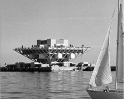 Black-and-white photo of a large elevated structure on a waterfront with a sailboat in the foreground