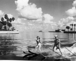 Two people water skiing on a calm bay, with palm trees and buildings visible along the shoreline in this vintage black-and-white photo