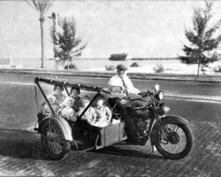 Man riding a motorcycle with a large sidecar carrying several young children, in a vintage black-and-white photo
