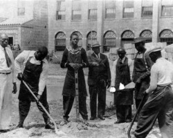 Black and white photo of a group of Black men working and standing near a brick building, some holding shovels and tools outdoors