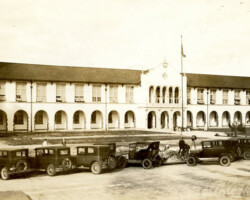 Historic sepia photo of a large Spanish Colonial Revival building with arched colonnades and vintage automobiles parked in front