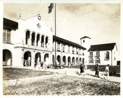 Vintage black-and-white photo of a Spanish Colonial Revival style building with arched colonnades and an American flag, with people walking outside