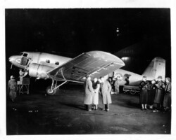 Passengers stand near an Eastern Airlines DC-3 aircraft at Albert Whitted Airport, c. 1930s. Workers for the Eastern Air Lines place their baggage in the plane NC 13737. The ladies are dressed in furs and small hats. The men wear trench coats and fedoras. A building in the background says "Mail" on the front, but it being dark, the rest of the sign is hard to read. Passengers stand near an Eastern Airlines DC-3 aircraft at Albert Whitted Airport, c. 1930s. Workers for the Eastern Air Lines place their baggage in the plane NC 13737. The ladies are dressed in furs and small hats. The men wear trench coats and fedoras. A building in the background says "Mail" on the front, but it being dark, the rest of the sign is hard to read.