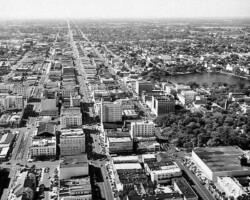 Looking west along Central Ave. from 3rd St, c. 1955. Gas Plant and Webb's City in the upper left. Mirror Lake and Williams Park in the upper right. Atlantic Coast Line along 1st Ave. S. Black and white aerial view of a mid-20th century American city with a grid street layout, downtown buildings, and a lake visible to the right